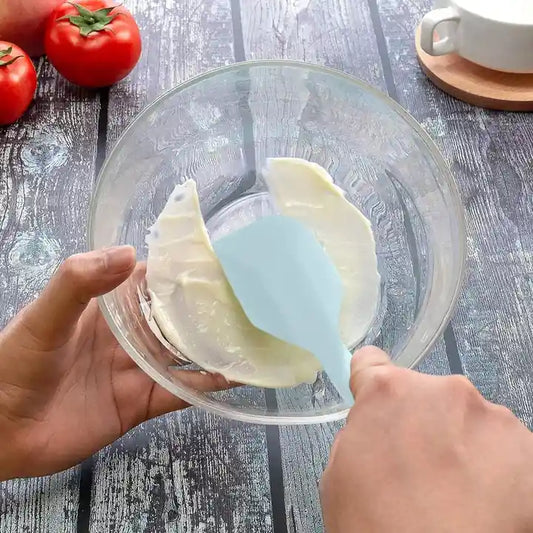 Person using a blue spatula to mix ingredients in a glass bowl on a wooden surface with tomatoes and a cup in the background.